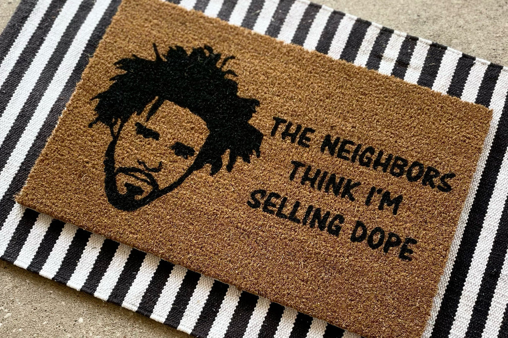 The Doormat District doormat with the text The Neighbors Think I'm Selling Dope with an outline of J. Cole's face next to it. 