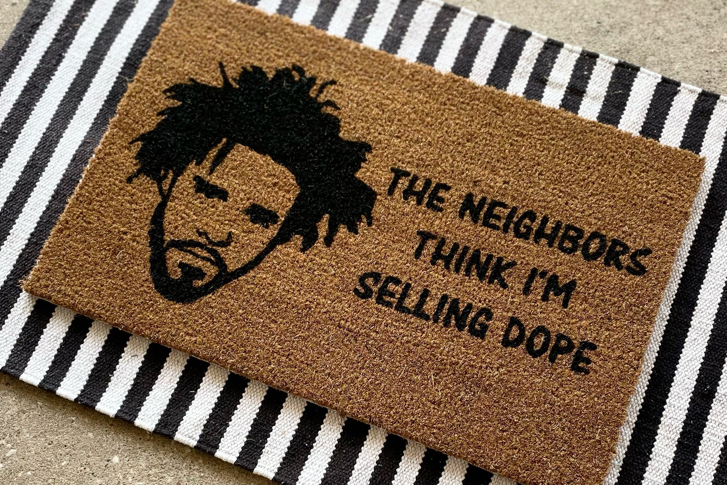 The Doormat District doormat with the text The Neighbors Think I'm Selling Dope with an outline of J. Cole's face next to it. 