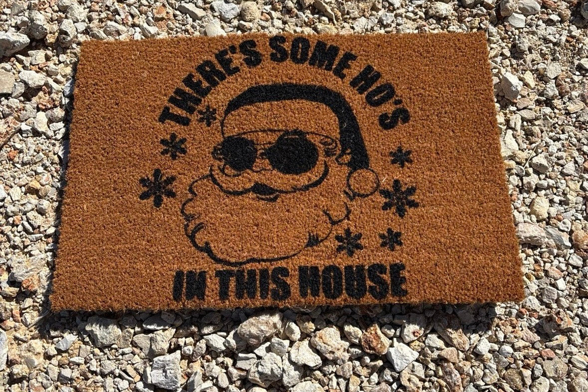 The Doormat District with a humorous design of a Santa Claus face and text that reads, "There's Some Ho's In This House"